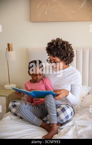 African american mother and daughter reading children book on bed with headboard, lamp Stock Photo