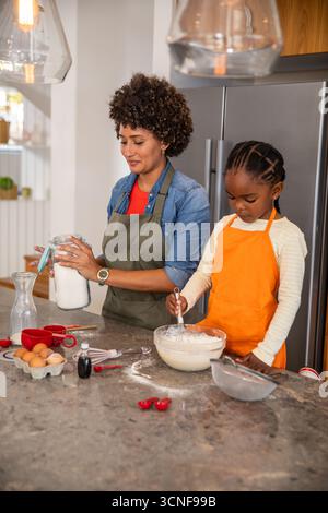 Diverse mother with daughter whisking batter at kitchen island in aprons with mixing bowl and jar Stock Photo