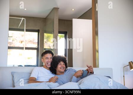 Diverse male partners snuggling under gray duvet on light sofa holding ceramic mug by window Stock Photo