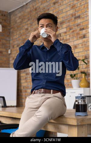 Asian man drinking coffee from white ceramic mug beside french press on desk in workspace Stock Photo