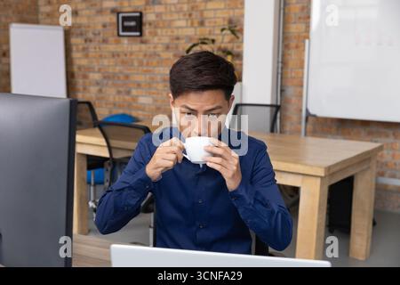 Asian man sitting at modern office desk facing computer while sipping coffee from white ceramic cup Stock Photo