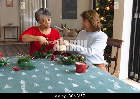 Diverse senior female friends cutting twine and arranging wreath at living room table with red mug Stock Photo
