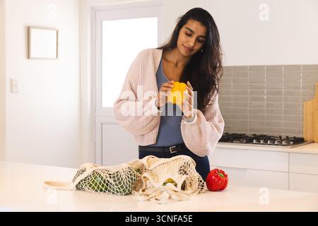 Asian woman holding yellow bell pepper and inspecting mesh bags and red pepper on island countertop Stock Photo