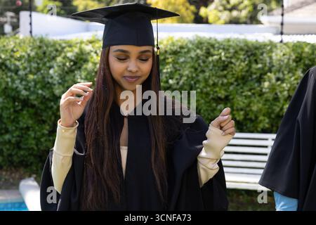 Female student wearing black gown adjusting mortarboard cap and standing on campus by bench Stock Photo