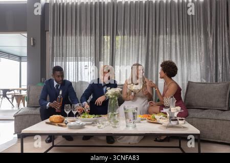 Bride and groom with friends sitting on sofa at lounge sharing snacks sipping wine Stock Photo