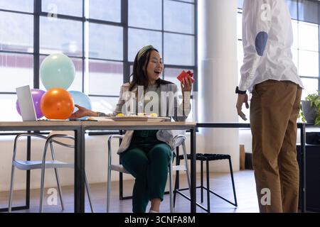 Diverse coworkers celebrating in office chinese woman holding red gift box by cake, balloons Stock Photo