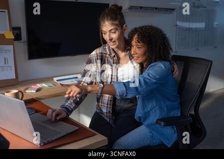 Diverse female coworkers sitting at wooden office desk collaborating on laptop, reviewing charts Stock Photo