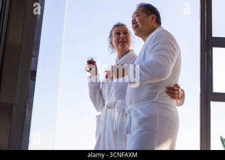 Senior couple standing by glass window in spa wearing bathrobes holding glass mugs with tea Stock Photo