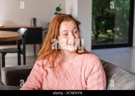 Woman sitting on gray sofa smiling while looking right in living room with glass window Stock Photo