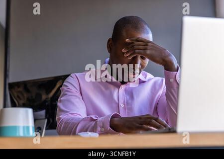African american man rubbing forehead, sitting at desk in office with laptop monitor Stock Photo