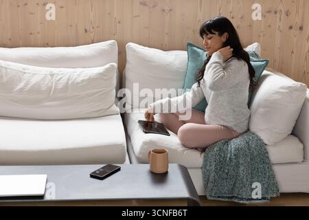 Hispanic woman sitting on sofa at home touching hair while using tablet next to mug, copy space Stock Photo