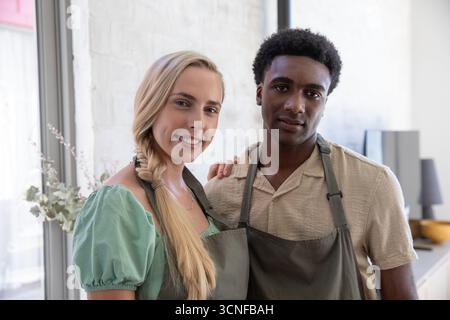 Diverse couple wearing grey aprons standing in modern kitchen near glass vase and bowl Stock Photo