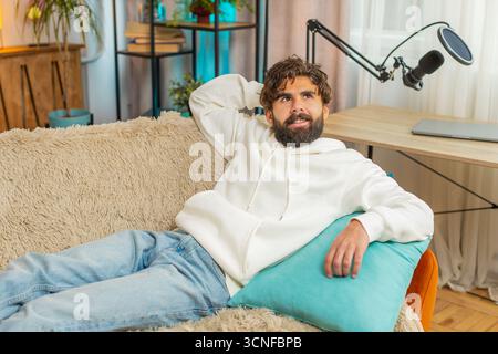 Tired Indian handsome young man enjoy relaxing on home sofa in living room. Happy guy lying on couch resting napping after hard working day, closed his eyes, put his hand behind his head, stress free. Stock Photo
