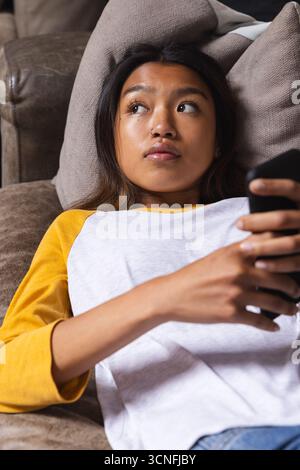 Asian teenage female reclining on brown leather couch in living room holding dark smartphone Stock Photo