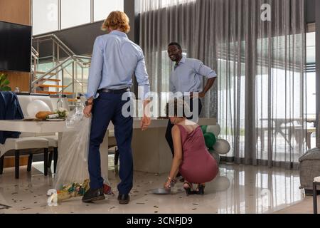 Diverse friends sweeping gold confetti with dustpan, packing decorations into bag in dining area Stock Photo