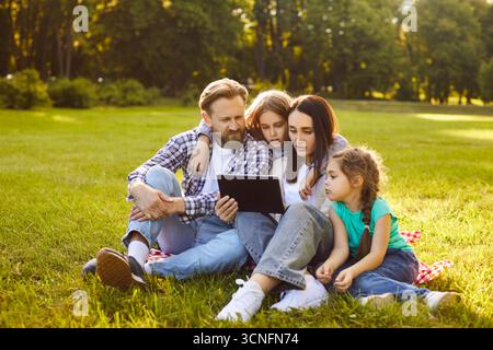 Happy family using tablet, reading digital book and sitting together on green grass of summer park Stock Photo