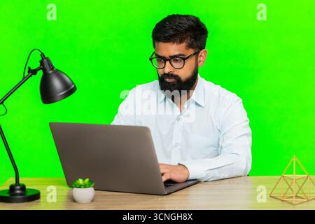 Indian man types on laptop with concentrated expression, showing remote work or studying in a focused manner. Arabian guy isolated on green chroma key background expressing productivity and dedication Stock Photo