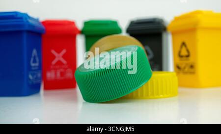 Plastic bottle caps in focus with garbage bins in the background representing the global micro plastics pollution crisis and recycling challenges. Con Stock Photo