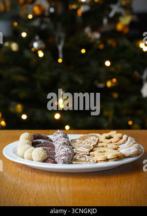 Gingerbread, Linzer and other types of Christmas cookies on a white plate standing on wooden table, a decorated glowing Christmas tree in the backgrou Stock Photo