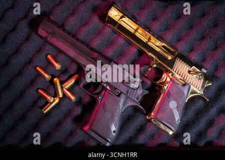A pair of powerful semi-automatic handguns, one gold-plated and one black, displayed with ammunition on a dark foam surface. Stock Photo