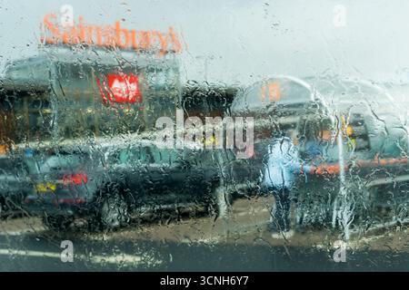 Supermarket shopping in the rain. Shopper returns trolley in a Sainsbury's car park during a rainstorm - viewed through a wet window. Stock Photo