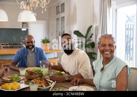 Diverse family sitting around wooden dining table in home kitchen sharing roasted chicken and salad Stock Photo