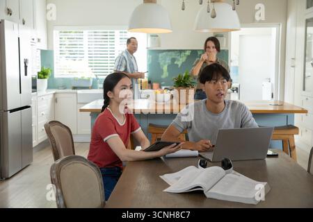 Asian siblings studying together using laptop, tablet, textbook at home kitchen dining table Stock Photo