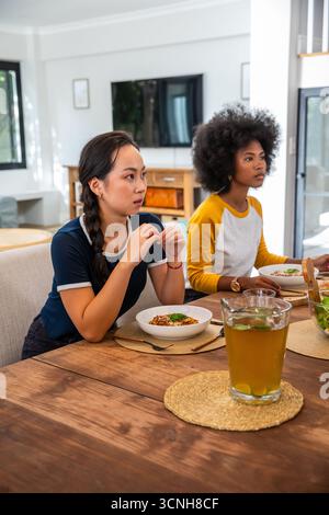 Diverse female friends sitting with pasta bowls and woven placemats at dining table at home Stock Photo