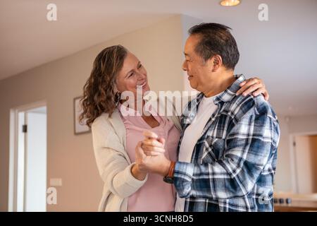 Diverse senior couple dancing in living room under recessed lights with picture Stock Photo