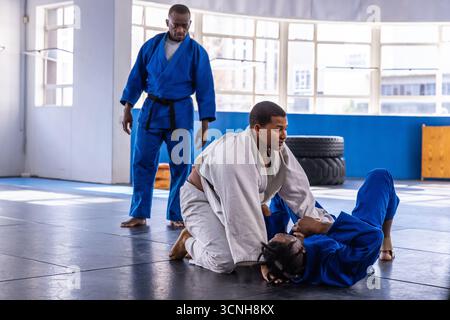 Diverse male martial-arts instructor supervising grappling in gis with two students on mat Stock Photo