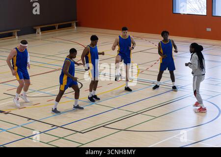 Diverse male basketball team wearing blue jerseys warming up on court with coach in tracksuit Stock Photo