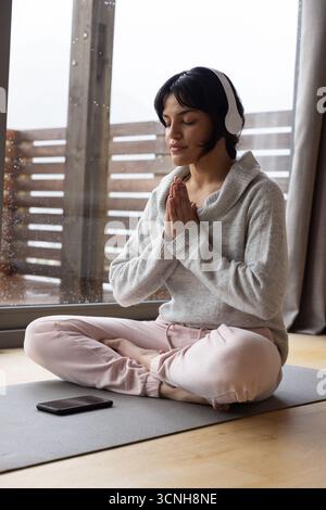 Asian woman sitting in loungewear on yoga mat by door meditating with smartphone, headphones Stock Photo