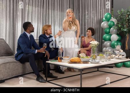 Diverse wedding party raising rosé glasses at reception lounge, bride in gown, bread on table Stock Photo