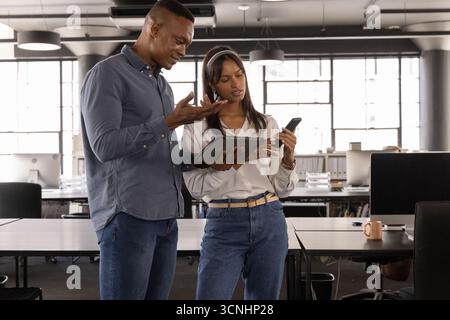 Diverse coworkers standing in office using tablet and smartphone near desks with monitors Stock Photo