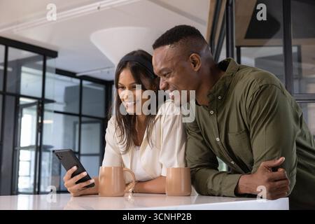Diverse coworkers leaning on white countertop in break room sharing smartphone near two tan mugs Stock Photo