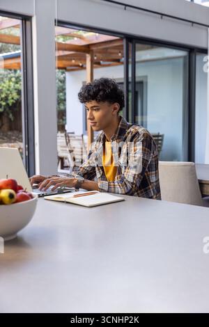 Asian male student typing on laptop at kitchen with notebook and apples, copy space Stock Photo