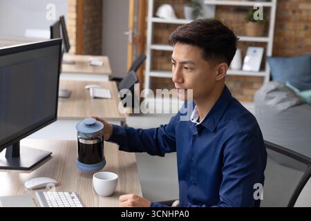 Asian man brewing coffee with french press at office desk with monitor, mug Stock Photo