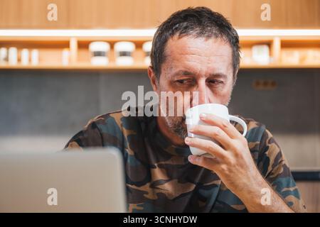 Mature adult caucasian male wearing camouflage army shirt drinking coffee and using laptop computer at home, selective focus Stock Photo