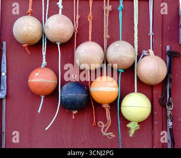Vintage fishing floats of various colors are neatly arranged and hanging from ropes on a rustic red wooden wall, showcasing a coastal village’s mariti Stock Photo