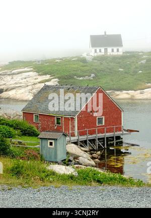 A serene coastal scene at Peggy's Cove, Nova Scotia captures a red boathouse nestled by the water, surrounded by fog. Old houses dot the landscape Stock Photo