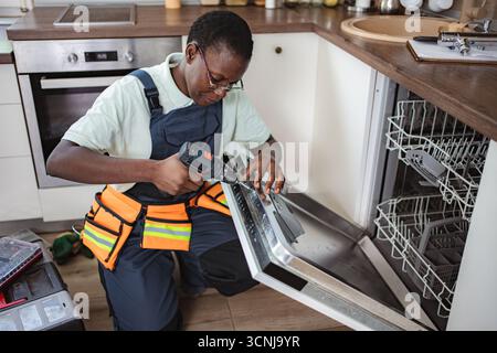Technician repairing dishwasher using electric screwdriver in kitchen Stock Photo