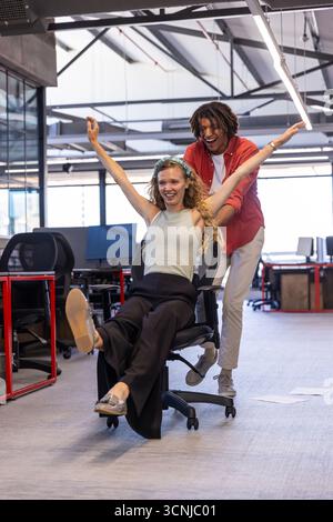 Diverse coworkers pushing rolling office chair and laughing together in modern open-plan office Stock Photo