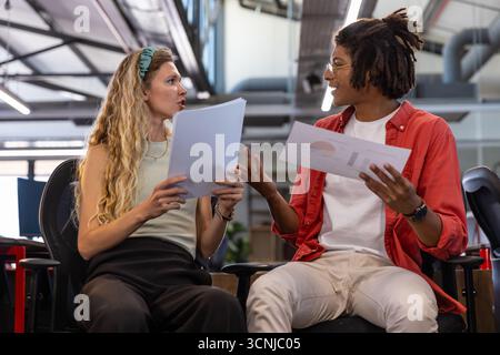 Diverse coworkers sitting in rolling chairs at open office discussing printed charts and graphs Stock Photo