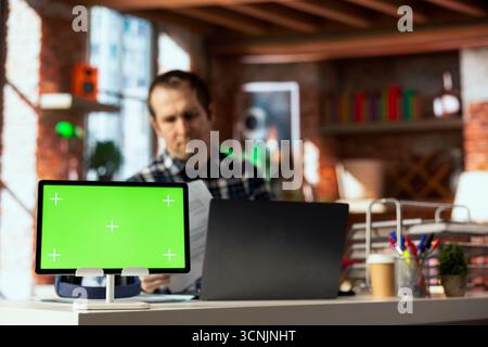 Green screen tablet in front of man in home office crosschecking information between laptop screen and paper files. Close up of chroma key device next to employee checking documents Stock Photo