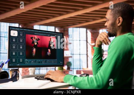 Smiling man in apartment workspace using AI software on computer display turning text prompt into photos. Upbeat african american person in home office uses text to image generative models on PC Stock Photo