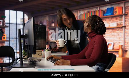 Exhausted AI startup employee rubbing forehead while analyzing datasets on computer, receiving coffee from project CTO manager to get energy and continue coding, camera B Stock Photo