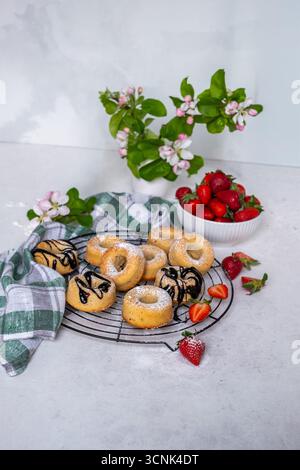 Delicious donuts cooling on a wire rack, featuring chocolate drizzle and powdered sugar, with fresh strawberries and greenery creating a vibrant desse Stock Photo