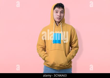 A casual young man wears a mustard-yellow hoodie and stands against a soft pink backdrop, Stock Photo