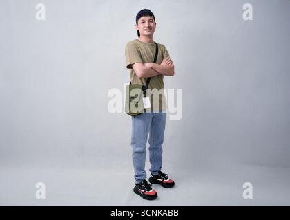 A young man stands with arms crossed, wearing a beige t-shirt, blue jeans, cap, and an olive crossbody bag in studio background Stock Photo