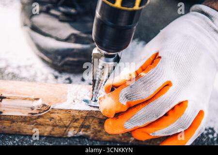 Gloved hand expertly guides a drill with a step bit, creating a precise hole in a wooden plank. Close-up captures focused craftsmanship and DIY Stock Photo